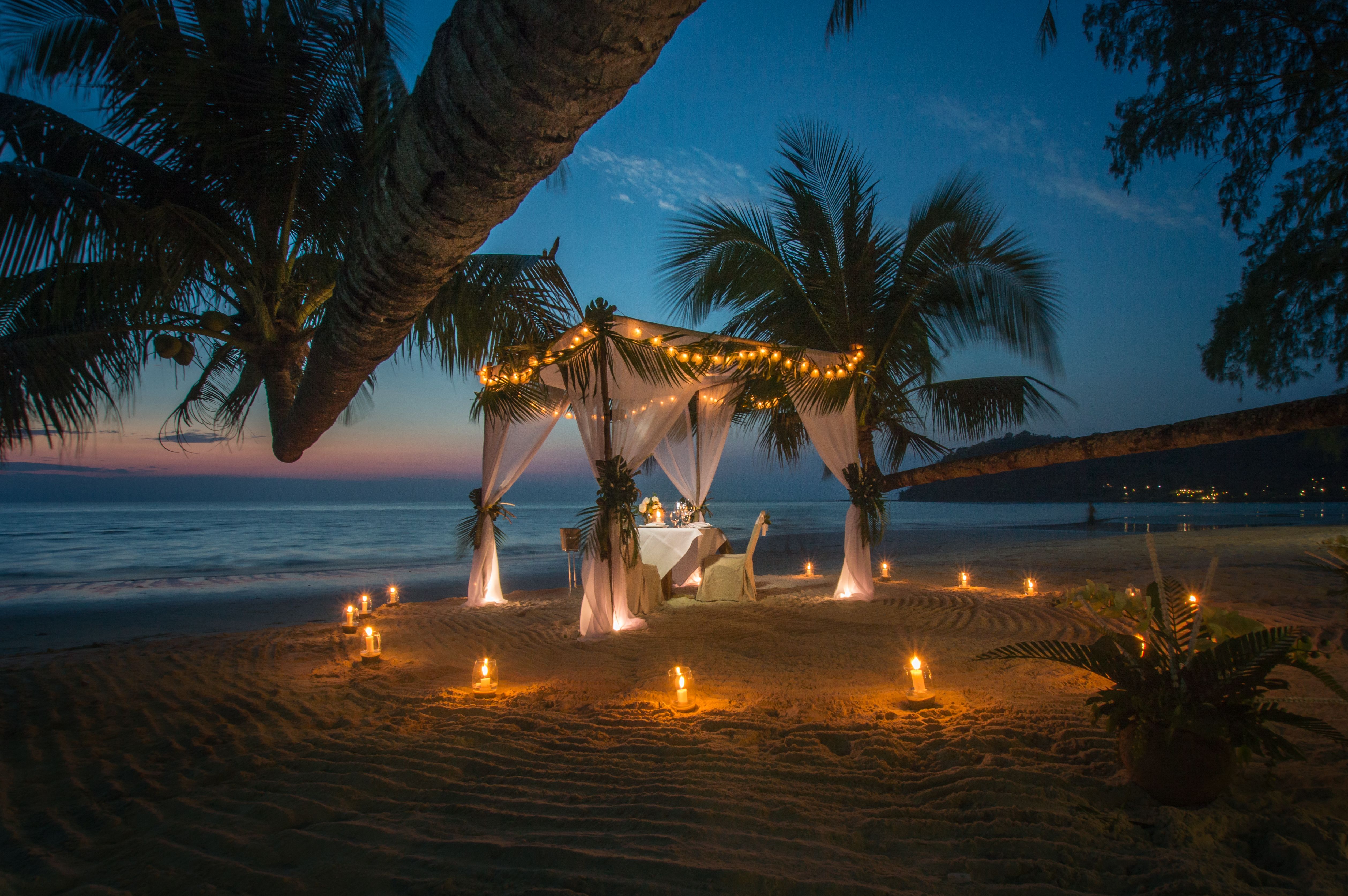 Candlelit dinner table beneath a draped canopy on the beach at dusk